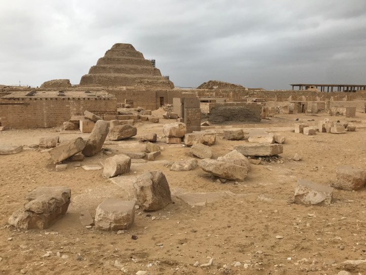 A sandy field covered in rocks with the Step Pyramid in the background